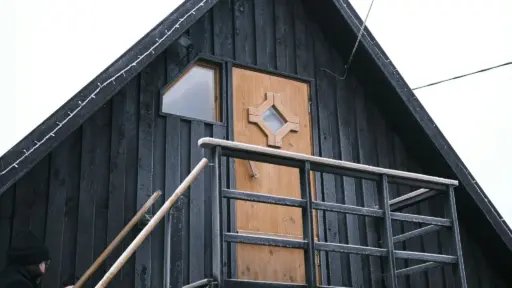Black wooden cabin exterior with person climbing snow-dusted staircase and geometric door design