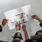 Two people examining architectural floor plan in open, unfinished room with concrete flooring
