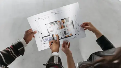 Two people examining architectural floor plan in open, unfinished room with concrete flooring