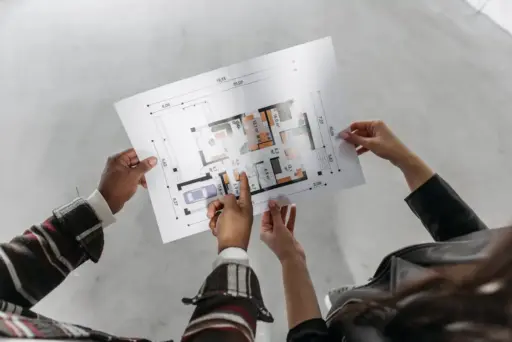 Two people examining architectural floor plan in open, unfinished room with concrete flooring