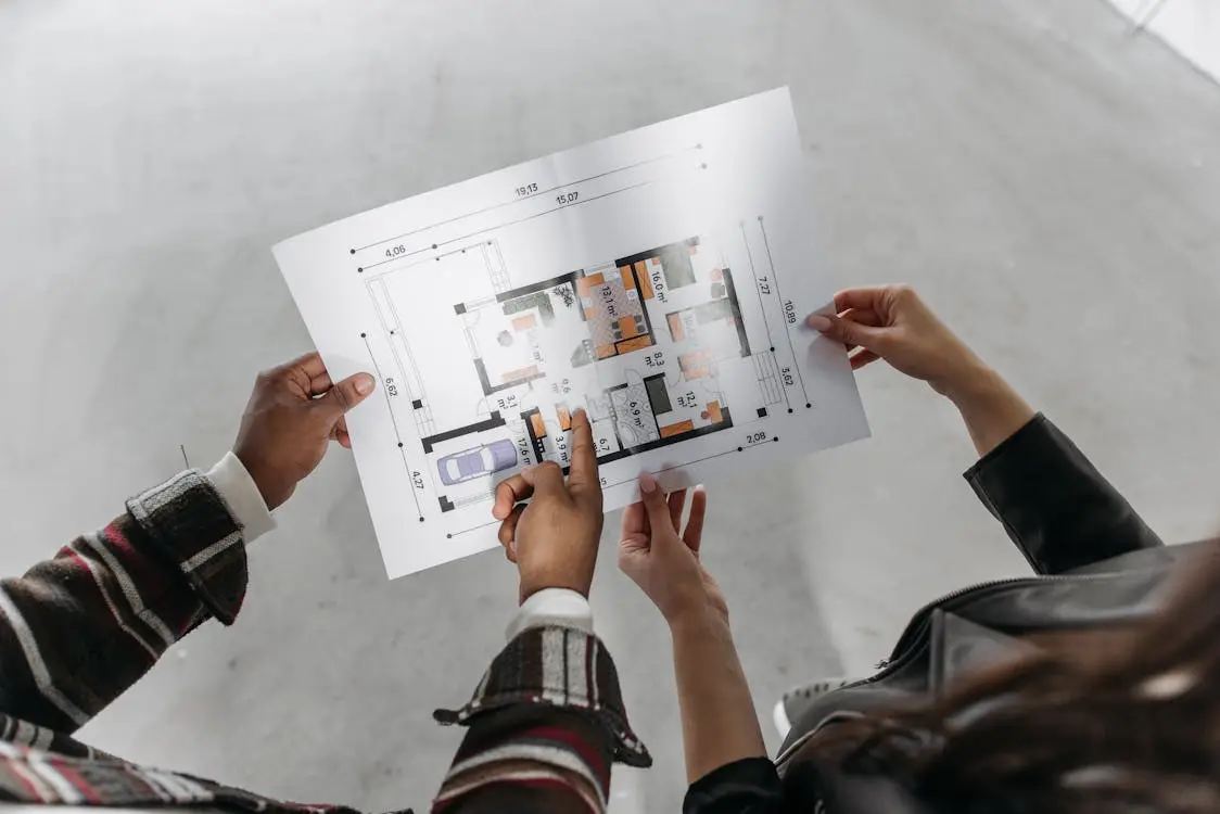 Two people examining architectural floor plan in open, unfinished room with concrete flooring