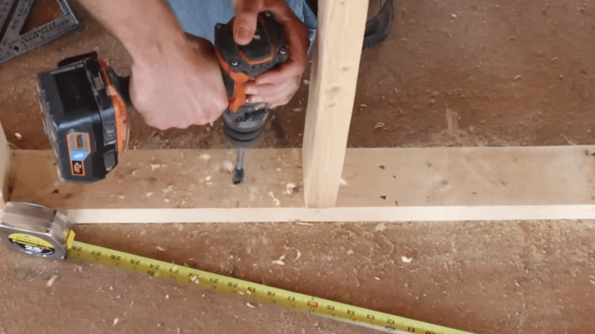 worker drilling through wooden stud base plate with drill while tape measure lies extended across dusty floor