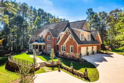 Large brick house with gabled roof and manicured lawn in wooded suburban setting