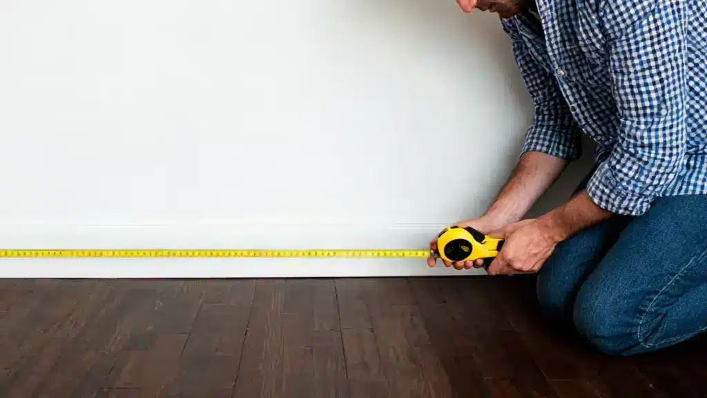Man in plaid shirt kneeling to measure the baseboard length on dark wood flooring against a white wall using a yellow tape measure