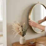 a person adjusting a round brass mirror over a wooden console table with a measuring tape and pampas grass nearby
