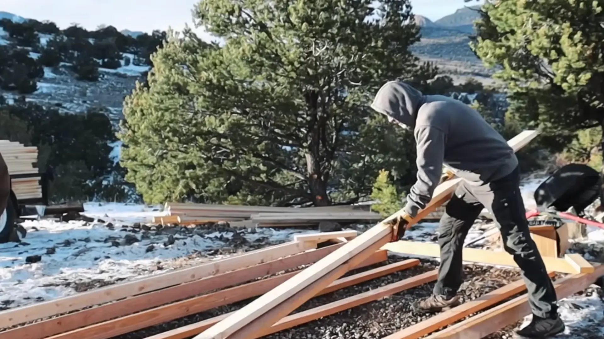 a person lifting and arranging wooden lumber boards outdoors while building a DIY sauna structure from scratch