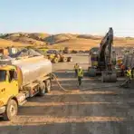 Construction site with yellow trucks and excavator in hilly landscape under clear blue sky