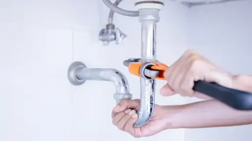 Hands using wrench to tighten pipe under sink in bright bathroom setting
