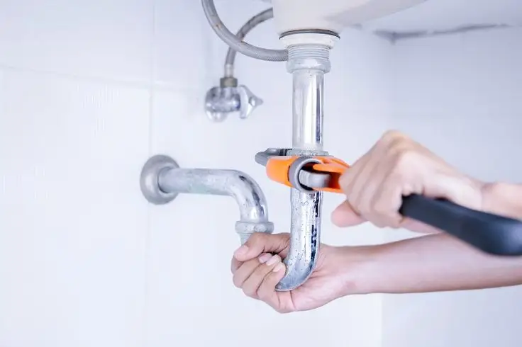 Hands using wrench to tighten pipe under sink in bright bathroom setting