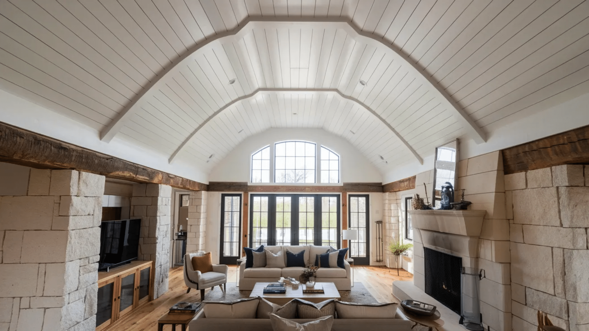 bright living room with vaulted wood plank ceiling large arched window stone fireplace sofas and glass doors opening to outdoor view