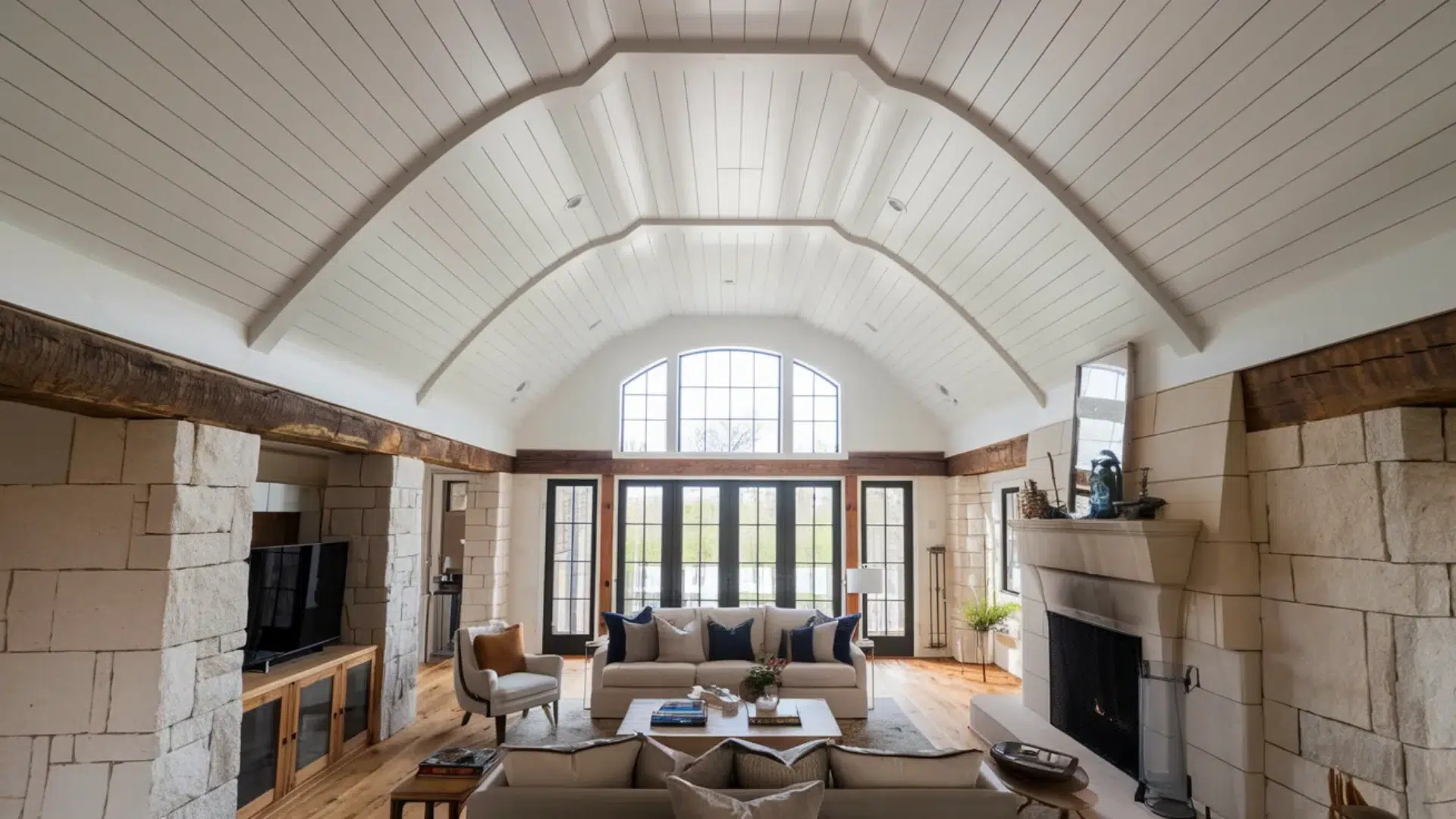 bright living room with vaulted wood plank ceiling large arched window stone fireplace sofas and glass doors opening to outdoor view