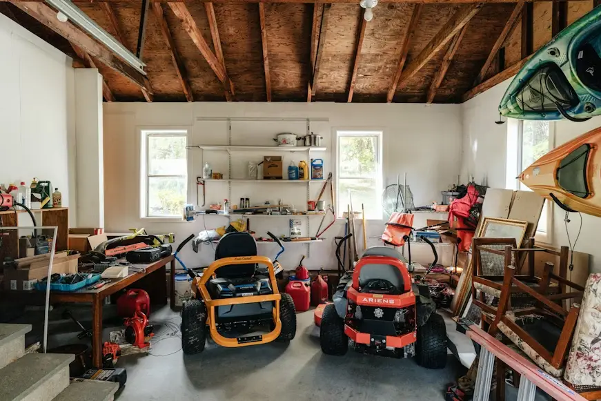 Lawnmowers and kayaks in cluttered garage with wooden ceiling and shelves filled with tools