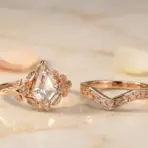 Rose gold engagement ring set with intricate leaf designs on marble surface with scattered petals