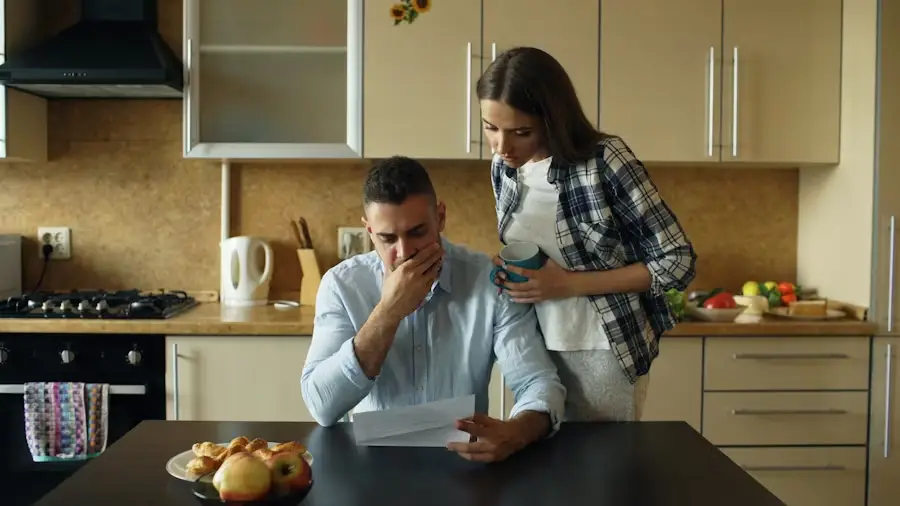 Man reading a document at kitchen table with woman holding mug nearby