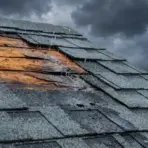 Damaged roof with missing shingles and exposed wood during rainy weather