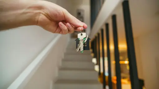 Hand holding house keys in modern hallway with staircase in soft lighting