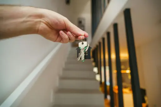 Hand holding house keys in modern hallway with staircase in soft lighting