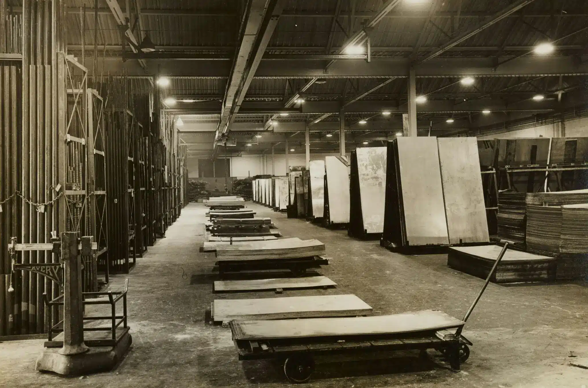 Industrial warehouse interior with metal sheets stacked and trolleys on concrete floor under fluorescent lights