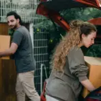Two people loading cardboard boxes into the trunk of a red car outdoors