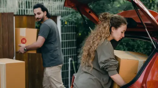 Two people loading cardboard boxes into the trunk of a red car outdoors