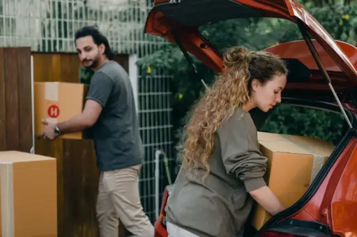 Two people loading cardboard boxes into the trunk of a red car outdoors