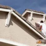 Person painting house shutters on a beige exterior under clear blue sky
