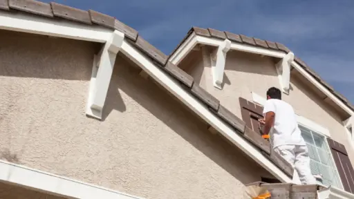 Person painting house shutters on a beige exterior under clear blue sky