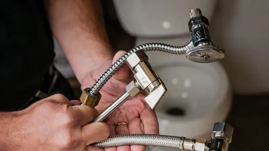 Hands holding plumbing pipes and valves in front of a toilet in a bathroom setting