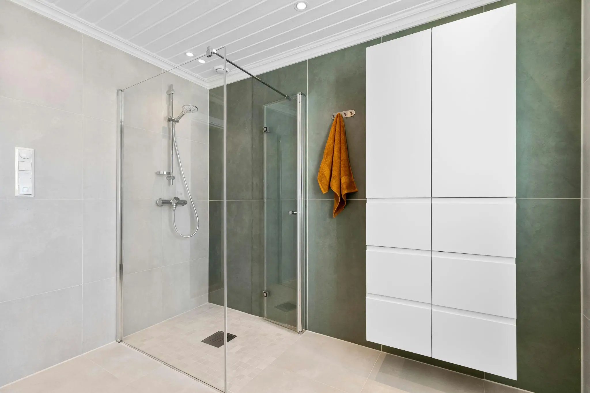 Modern bathroom with glass shower enclosure, white storage cabinets, and orange towel on green wall