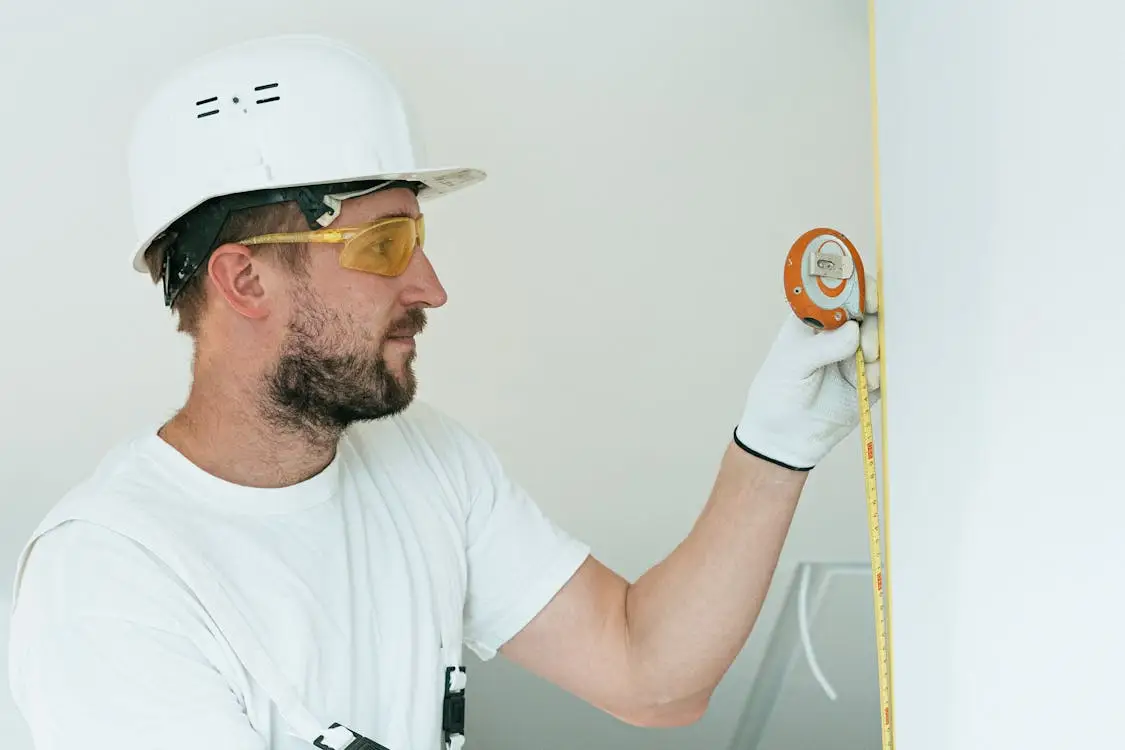 Construction worker measuring wall indoors with yellow tape measure and wearing white hard hat