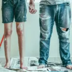 Barefoot couple holding paintbrushes standing on newspapers in room with white walls and paint stains