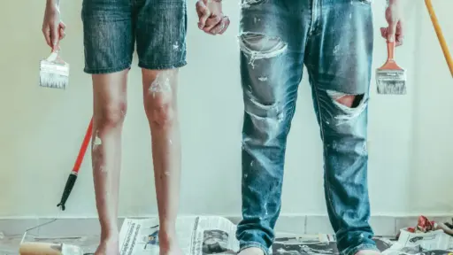 Barefoot couple holding paintbrushes standing on newspapers in room with white walls and paint stains