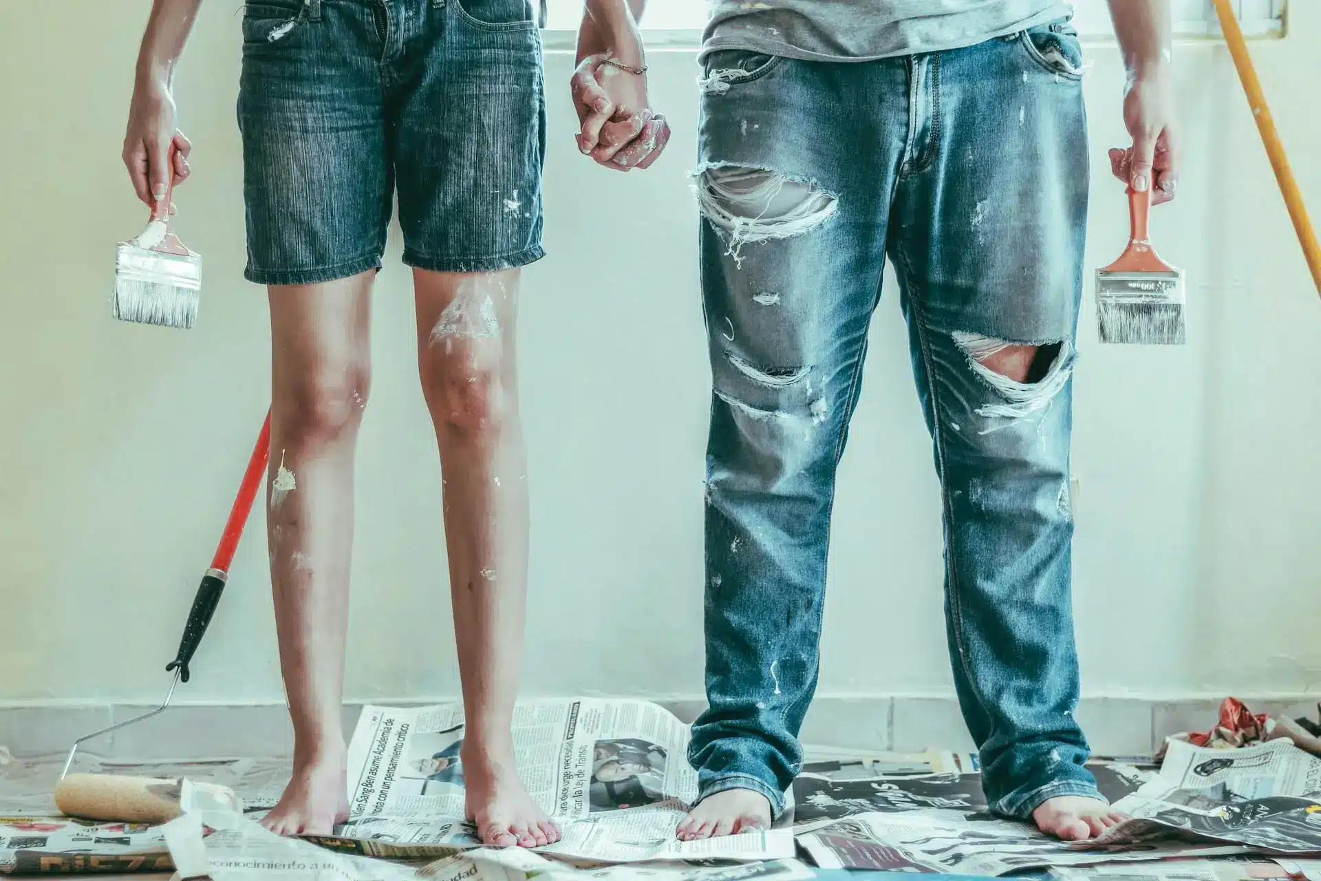 Barefoot couple holding paintbrushes standing on newspapers in room with white walls and paint stains