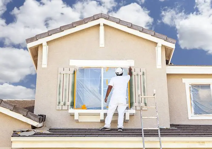 Painter in white uniform working on exterior house shutters under blue sky with clouds