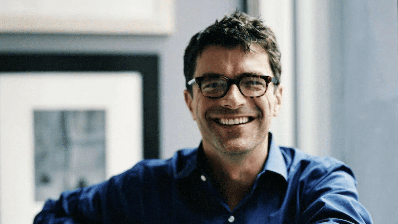 thomas o'brien wearing glasses and a blue shirt leaning on a railing indoors, with framed artwork visible in the softly lit background