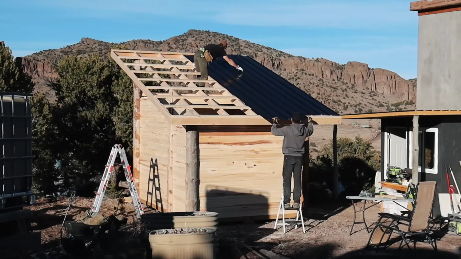 two people installing a steep-angled metal roof over a completed aspen tongue and groove sauna structure with wood framing.