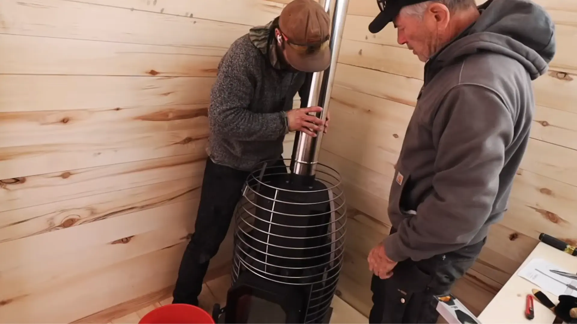 two people installing a wood burning stove with a stainless steel stove pipe inside a sauna with aspen wood walls
