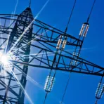 Transmission tower silhouette against bright blue sky with sunlight radiating through steel framework
