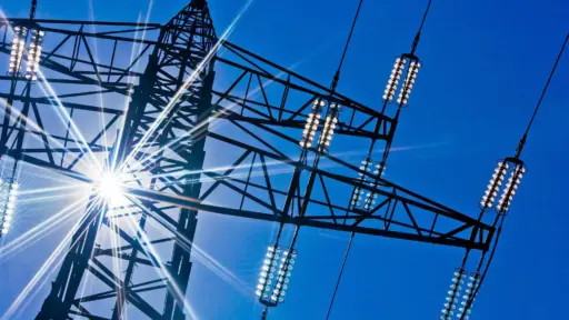 Transmission tower silhouette against bright blue sky with sunlight radiating through steel framework