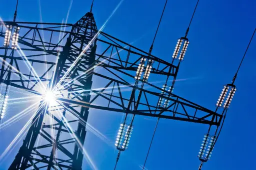 Transmission tower silhouette against bright blue sky with sunlight radiating through steel framework