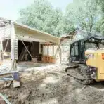 Excavator demolishing a house exterior in a residential area under clear skies