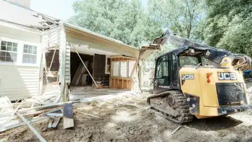 Excavator demolishing a house exterior in a residential area under clear skies