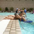 Group of women practicing swimming strokes in an outdoor pool with potted plants nearby
