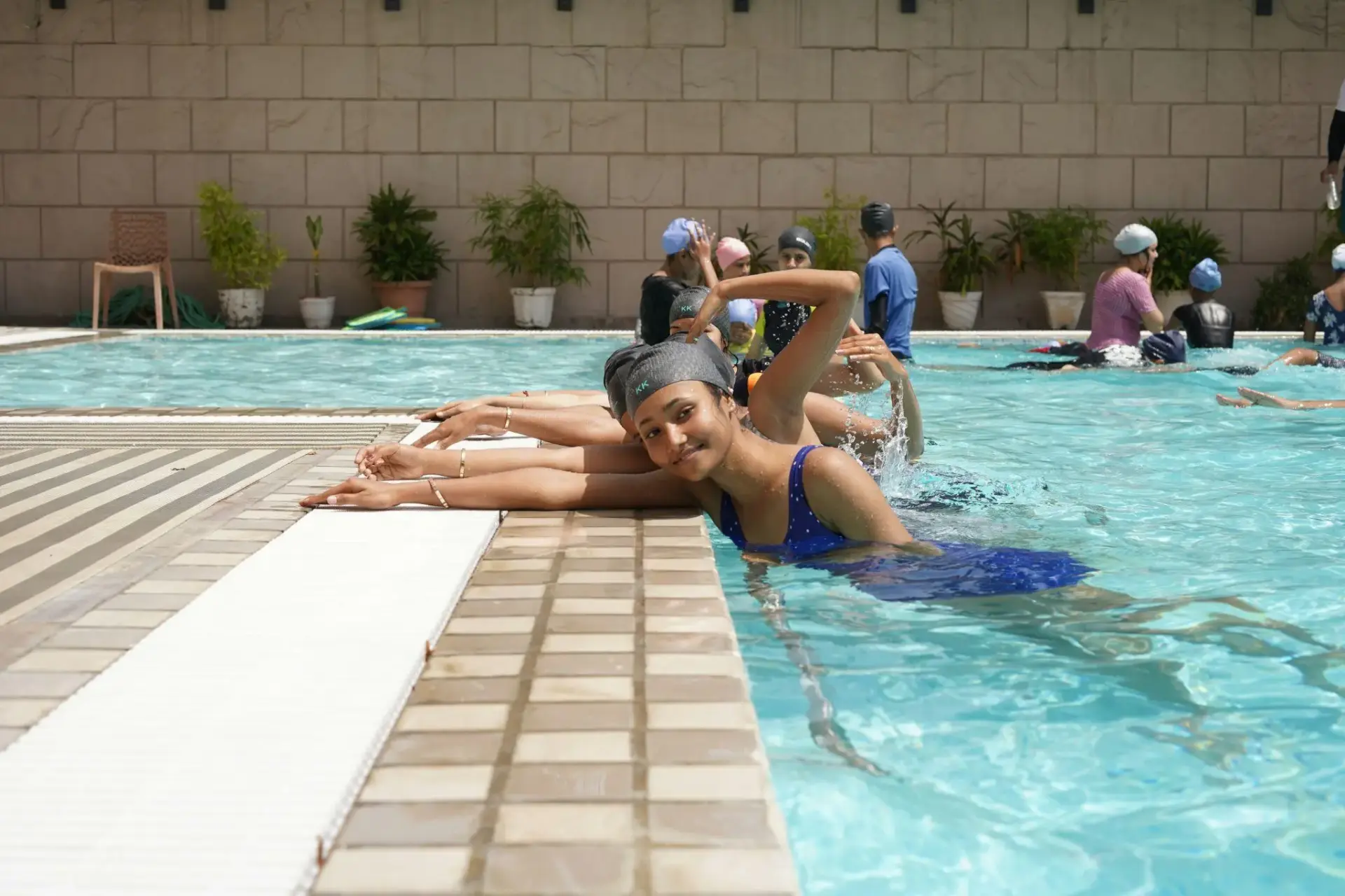 Group of women practicing swimming strokes in an outdoor pool with potted plants nearby
