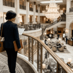 woman in business attire standing on an upper-level balcony, reviewing documents while overlooking an elegant hotel lobby with chandeliers and guests below