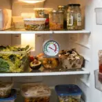 Refrigerator interior with various food containers, produce, jars, and a hanging thermometer