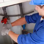 Worker in blue uniform using wrench on pipes under sink in utility room