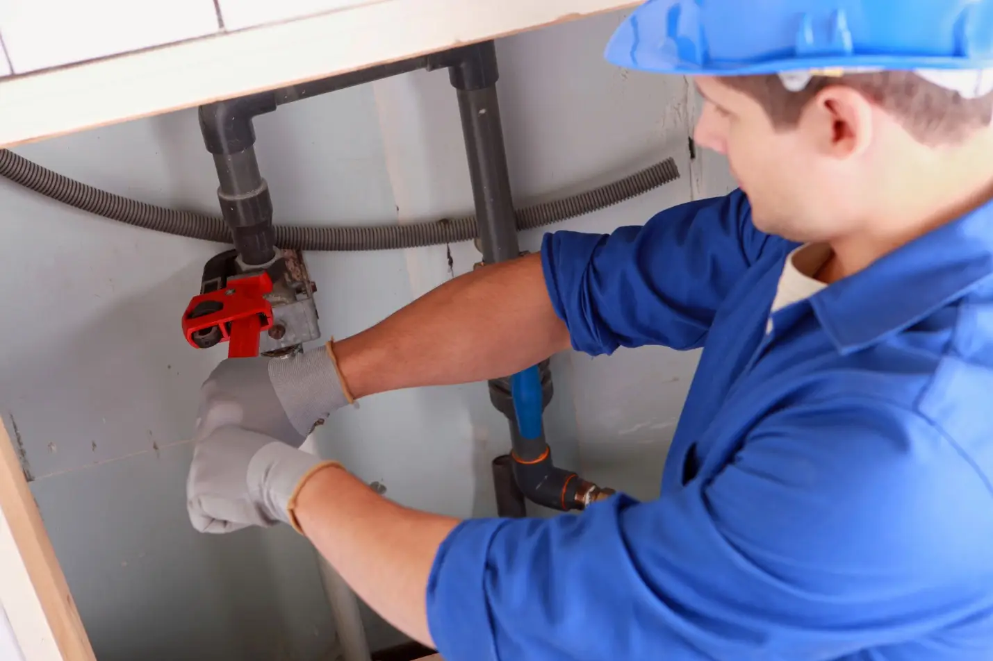 Worker in blue uniform using wrench on pipes under sink in utility room