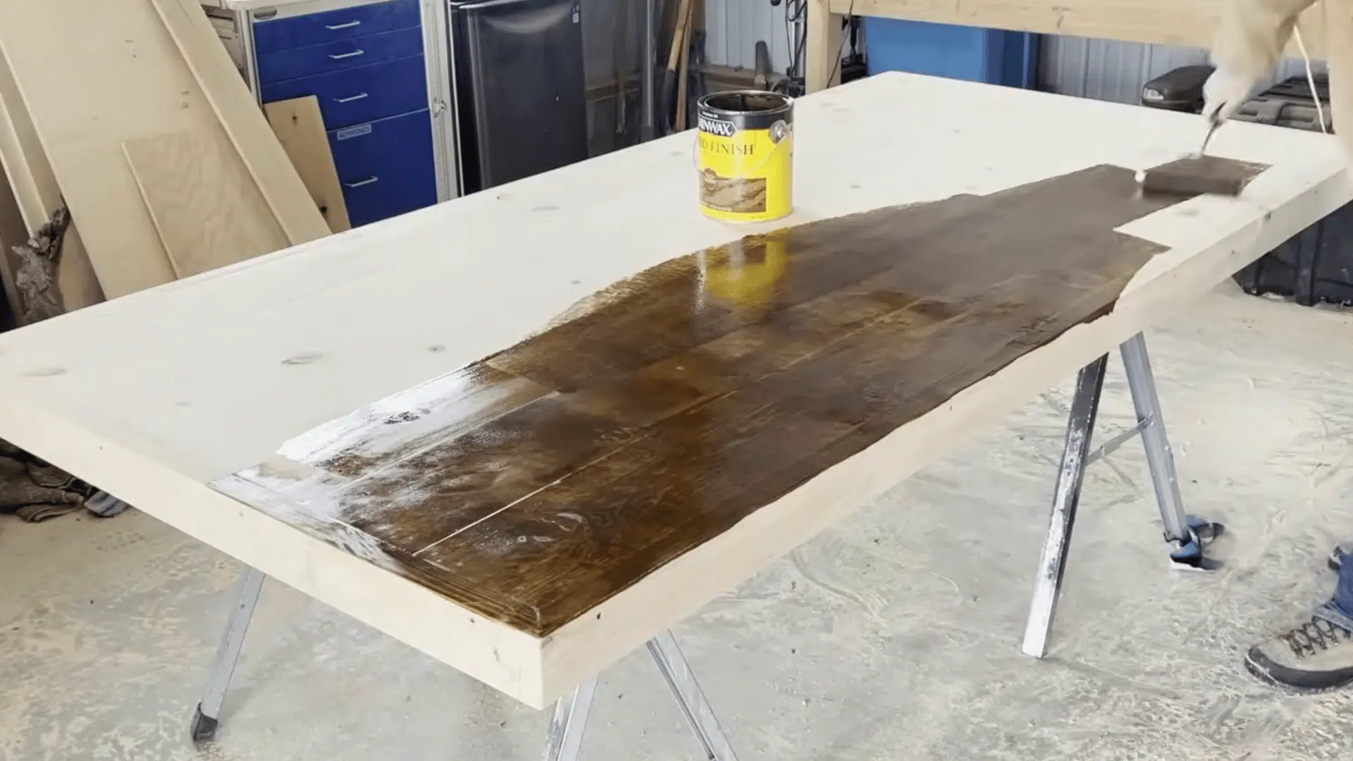 a woodworker applying dark wood stain across a large tabletop creating a rich finish in a workshop