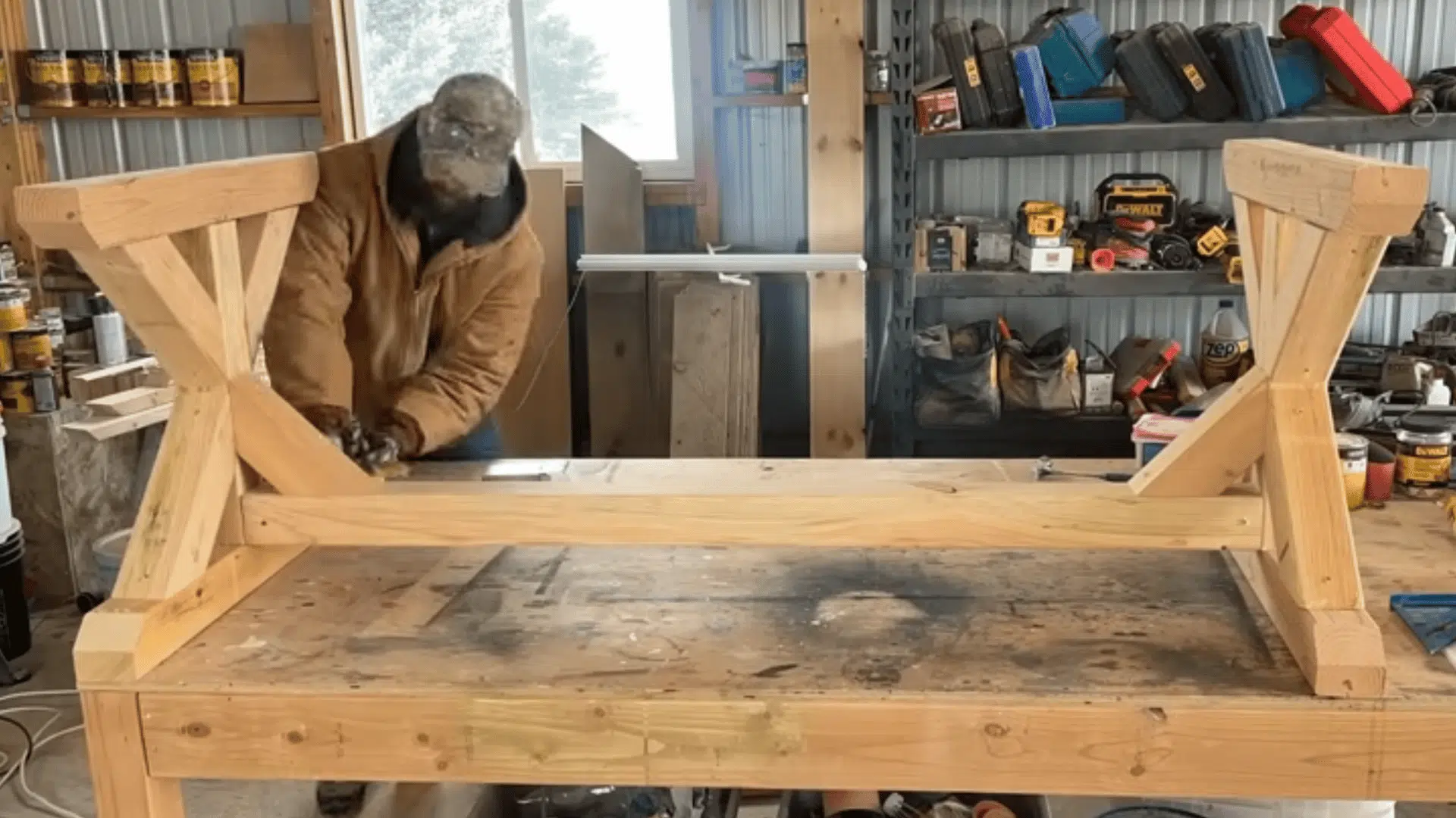 a woodworker measuring and adjusting a wooden table base frame on a workbench surrounded by tools and supplies